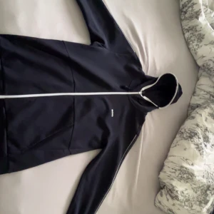 Svart hoodiejacka från BOSS med dragkedja - Säljer en svart hoodiejacka från BOSS med vit dragkedja och diskret logga på bröstet. Koftan har huva, långa ärmar och fickor framtill. Perfekt för en sportig och stilren look.