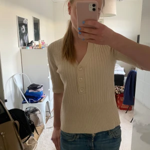Beige ribbad topp med knappar - Säljer en beige ribbad topp med v-ringning och trekvartsärm. Toppen har dekorativa knappar framtill och är i ett mjukt, stretchigt material. Perfekt till jeans eller kjol för en enkel och stilren look.