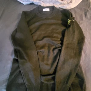 Svart stickad tröja från Stone Island - Säljer en svart stickad tröja från Stone Island med klassisk rund halsringning och ribbade muddar. Tröjan har den ikoniska Stone Island-loggan på ärmen och är perfekt för dig som gillar stilrena plagg med premiumkänsla.