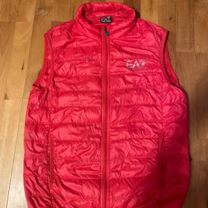 Röd dunväst från Emporio Armani EA7 - Snygg röd dunväst från Emporio Armani EA7 med dragkedja framtill och logga på bröstet. Västen har en quiltad design och hög krage, perfekt för lager-på-lager-stil.