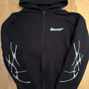 Svart hoodie med tryck från Upscale Studios - Svart hoodie med dragkedja från Upscale Studios. Har grått 'speed'-tryck på bröstet, stort rosa och grått tryck på ryggen samt gråa grafiska detaljer på ärmarna. Mjuk och skön med huva och fickor framtill. Knappt använd.