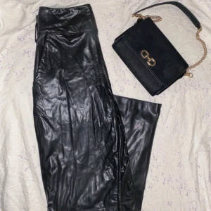 Svarta skinnbyxor från NA-KD (väska på köpet) - Säljer ett par svarta skinnbyxor från NA-KD med hög midja och raka ben. Byxorna har en snygg glansig finish och stängs med hake och dragkedja. Perfekta för dig som vill ha en trendig och edgy look.