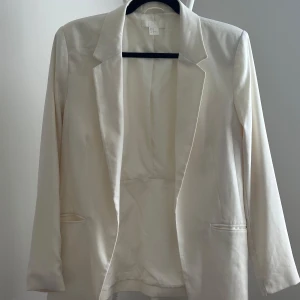 Vit kavaj från H&M - Snygg vit kavaj från H&M med klassisk krage och två fickor framtill. Långärmad modell med rak passform, perfekt för en stilren look. Tillverkad i ett slätt material som ger en elegant känsla. Storlek 40