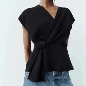 Svart omlottopp från Zara - Säljer en svart omlottopp från Zara med korta ärmar och snygg knytdetalj i midjan. Toppen har en v-ringad hals och en elegant, stilren design som passar perfekt till jeans eller kjol.