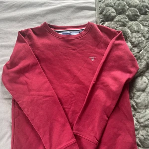 Röd sweatshirt från GANT - Säljer en röd sweatshirt från GANT med broderad logga på bröstet. Tröjan har rund halsringning med randig detalj på insidan och långa ärmar. Perfekt för dig som gillar klassisk och stilren design.