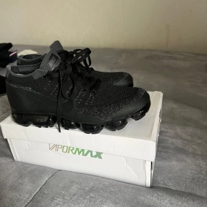Svarta Nike Vapormax sneakers - Säljer ett par svarta Nike Vapormax sneakers med stickad ovandel och klassisk snörning. Skorna har en tydlig, genomskinlig platt sula med luftbubblor för extra komfort och stil. Perfekta för dig som gillar sportig och modern look.