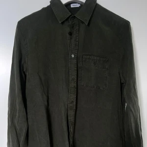 Mörkgrön skjorta från Filippa K - Stilren mörkgrön skjorta från Filippa K med klassisk krage och bröstficka. Skjortan har långa ärmar och knäppning framtill. Perfekt för en clean och enkel look. Storlek 48 = Storlek Small