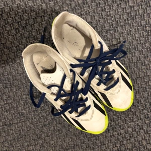Vita och neongula fotbollsskor från Adidas - Säljer ett par vita fotbollsskor från Adidas med neongula och svarta detaljer. Skorna har snörning och är perfekta för fotbollsspel på gräs. De har en iögonfallande design med neongula sulor och svarta ränder.