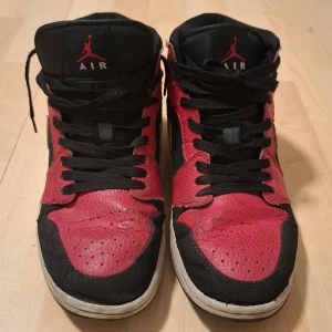 Svart och röd Nike Jordan 1 sneakers - Säljer ett par klassiska Nike Jordan 1 sneakers i svart och rött. Skorna har en hög modell med snörning och den ikoniska loggan på sidan. Perfekta för dig som vill ha en stilren och sportig look.