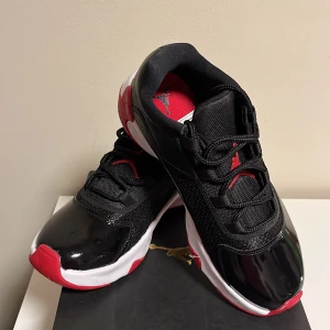 Air Jordan 11 CMFT Low (GS) - Snygga Air Jordan 11 CMFT Low sneakers i svart, vitt och rött. Skorna har en stilren design med snörning och en bekväm passform. Perfekta för både sport och vardag. (Oanvända)