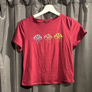 Röd t-shirt med svamptryck från Papaya - Säljer en cool röd t-shirt från Papaya med tre färgglada svampar tryckta på framsidan. T-shirten är kortärmad och gjord av en mjuk polyesterblandning. Skriv gärna om eventuella frågor.🍄