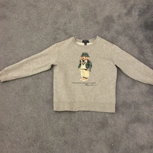 Grå tröja med Polo Bear från Ralph Lauren - Säljer en grå tröja från Ralph Lauren med det ikoniska Polo Bear-trycket på framsidan. Tröjan har långa ärmar och en rund halsringning. Ny pris på tröjan är 1300,säljer för 500. I utmärkt skick.