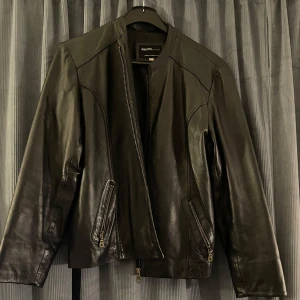 Svart skinnjacka  - Svart skinnjacka från med dragkedja framtill och två fickor med dragkedjor. Jackan har en stilren design med långa ärmar. Oklart vilken storlek jackan är i men ungefär M