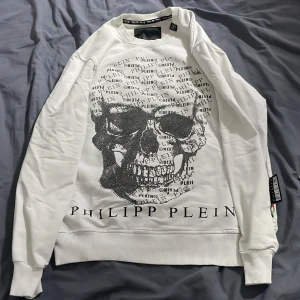 Vit tröja med dödskalle från Philipp Plein - Säljer en vit tröja från Philipp Plein med en stor dödskalle i svart och textdetaljer på framsidan. Tröjan har långa ärmar och ribbade muddar. Märkeslogga på baksidan och insidan av kragen. Passar m också