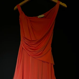 Orange/ rosa  klänning med drapering - Snygg orange/ rosa klänning med draperad design och asymmetrisk axel. Perfekt för en färgstark look. Den har inte används alls prisetikett finns kvar 