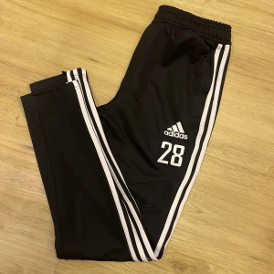 Svarta träningsbyxor från Adidas - Snygga svarta träningsbyxor från Adidas med vita ränder längs sidorna och nummer 28 tryckt på framsidan. Byxorna har elastisk midja och är perfekta för sport och träning.