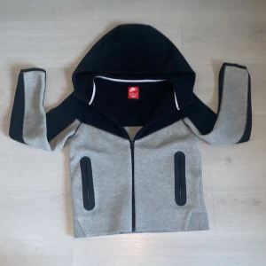Grå och svart hoodie från Nike - Säljer en snygg grå och svart hoodie från Nike med dragkedja och huva. Den har en modern design med svarta detaljer och två fickor framtill. Perfekt för en sportig look!