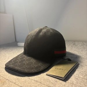 Svart Gucci keps - Storlek ”M” finns också! Snygg svart keps från Gucci med diskret mönster och klassiska ränder i rött och grönt på sidan. Perfekt för att ge din outfit en lyxig touch. Kepsen har en böjd skärm och justerbar passform. Storlek ”S”  