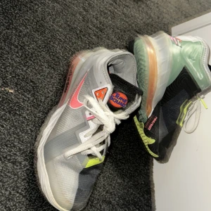 Nike sneakers med Space Jam-tema - Unika Nike sneakers med Space Jam-tema. Skorna har en mix av grå, vit och rosa färger med detaljer i neon. De har en synlig luftdämpning i sulan och snörning framtill. Innersulan har ett tryck av karaktärer från Space Jam.