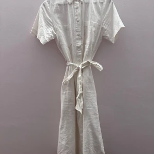 Vit klänning med knappar och knytskärp - Säljer en stilren vit klänning med korta ärmar och knytskärp i midjan. Klänningen har knappar framtill och är tillverkad i ett lätt och luftigt material, perfekt för varma dagar.