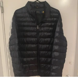  Ralph Lauren Jacka - Säljer en svart pufferjacka från Polo Ralph Lauren. Jackan har en quiltad design med dragkedja framtill. Perfekt för kyligare dagar med sin stilrena look och bekväma passform. 