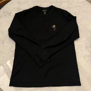 Svart långärmad tröja från Polo Ralph Lauren - Säljer en stilren svart långärmad tröja från Polo Ralph Lauren. Tröjan har en liten broderad detalj på bröstet som ger en extra touch. Perfekt för en avslappnad och snygg look.