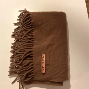 Brun halsduk från Acne Studios - Säljer en stilren brun halsduk från Acne Studios i 100% ull. Den har fransar i ändarna och en diskret logga. Perfekt för att hålla dig varm och snygg under kyliga dagar.