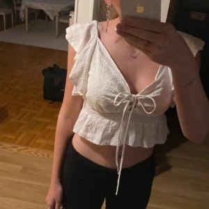 Vit blus från Wild Flower - Säljer en söt vit blus från Wild Flower med spetsdetaljer och volanger. Blusen är kortärmad och har en knytning framtill som ger en fin passform. Perfekt för varma dagar!