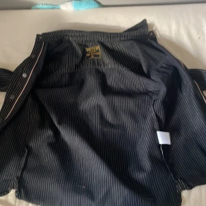 Svart randig skjorta från Pike Brothers - Säljer en stilren svart skjorta från Pike Brothers med vita ränder. Skjortan har knappar framtill och två bröstfickor. Perfekt för en klassisk look.