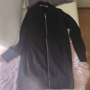 Svart lång cardigan från House of Lola - Säljer en stilren svart lång cardigan från House of Lola i storlek XS. Jackan har en dragkedja framtill och långa ärmar. Perfekt för en minimalistisk look.