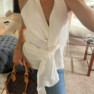 Vit omlottopp - Säljer en stilren vit omlottopp med knytning i midjan. Toppen är ärmlös och har en öppen rygg vilket ger en elegant look💖💖 lappen kvar och köpt för längesen så tror inte den säljs längre