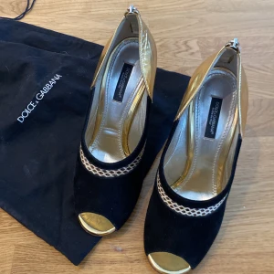 Svarta och guldiga klackskor från Dolce & Gabbana - Eleganta klackskor från Dolce & Gabbana i svart mocka med guldiga detaljer. Skorna är i svart mocka, och läder med en smal klack. Perfekta för en glamorös kväll.. 