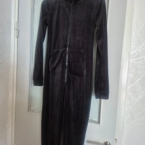 Svart onesie från Hunkemöller - Mysig svart onesie från Hunkemöller med dragkedja framtill och långa ärmar. Perfekt för kalla dagar när du vill hålla dig varm och bekväm. Den har en praktisk ficka framtill och är tillverkad i ett mjukt material.