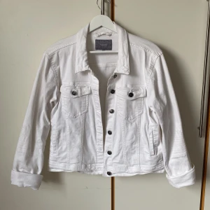 Vit jeansjacka från b.young - Snygg vit jeansjacka från b.young med klassisk design. Jackan har knappar framtill och två bröstfickor med knappdetaljer. Längd 56 cm. 