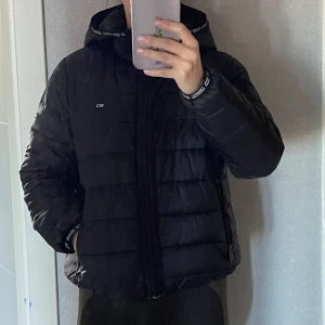 Svart jacka från Tommy Hilfiger - Säljer en svart jacka från Tommy Hilfiger med huva och dragkedja. Jackan har ett quiltat mönster och är perfekt för kyligare väder. Den har långa ärmar och två fickor med dragkedja framtill.