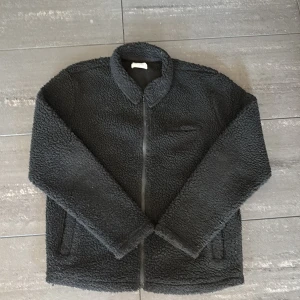 Svart teddyjacka från Studio Total - Mysig svart teddyjacka från Studio Total med dragkedja framtill. Jackan har en klassisk krage och två sidofickor. Bra för våren, relativt värmande