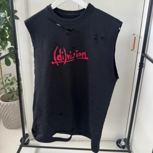 Svart ärmlös topp från Division - Säljer en cool svart ärmlös topp från Division med röd logga på framsidan. Toppen har en distressed look med flera slitningar för en edgy stil. Perfekt för dig som gillar en unik och avslappnad stil.