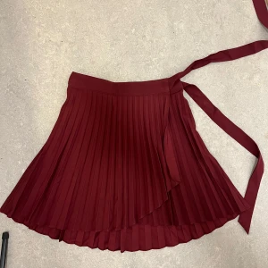 Vinröd plisserad kjol från NA-KD - Snygg vinröd plisserad kjol från NA-KD med omlott-design och knytband i midjan. Perfekt för att ge en elegant touch till din outfit. Kjolen har en kort längd och är tillverkad i ett lätt och bekvämt material. Mycket fint skick, använd max 1-2 gånger!