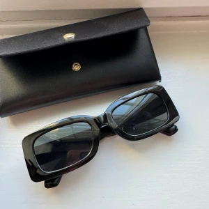 Svarta trendiga solglasögon - Snygga svarta solglasögon med rektangulära bågar. Perfekta för en stilren look. Kommer med ett matchande svart fodral.