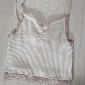 Vit ribbad linnetopp med spetsdetaljer - Säljer en söt vit ribbad linnetopp med smala axelband och spetsdetaljer i ljusrosa längs kanterna. Toppen har en v-ringad design och är perfekt för varma dagar.