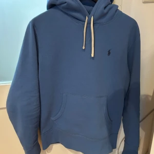 Blå hoodie från Ralph Lauren - Säljer en snygg blå hoodie från Ralph Lauren med klassisk känguruficka och justerbar huva med vita snören. Perfekt för en avslappnad stil.