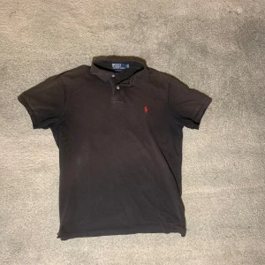 Svart pikétröja från Ralph Lauren - Säljer en klassisk svart pikétröja från Ralph Lauren med röd broderad logga på bröstet. Tröjan är i slim fit och tillverkad av 100% bomull. Perfekt för en stilren look!