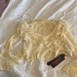 Gul spetsblus från Romwe - Säljer en söt gul spetsblus från Romwe med volangdetaljer och långa ärmar. Perfekt för en romantisk look. Blusen har en v-ringad design och knytband vid ärmsluten. Så somrig å fin.