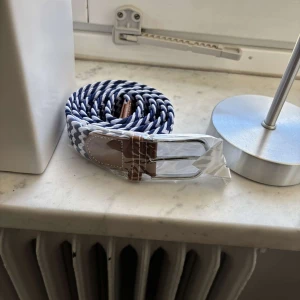 Flätat bälte - Snyggt randigt flätat bälte i blått och vitt med bruna skinn-detaljer. Perfekt för att ge en stilren touch till din outfit. Bältet har en klassisk design med ett spänne i silverfärg. 105 cm långt och elastiskt, passar därför alla.