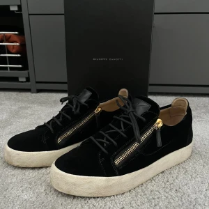 Svarta sneakers från Giuseppe Zanotti - Snygga svarta sneakers i mocka från Giuseppe Zanotti med guldfärgade dragkedjor på sidorna. Skorna har en vit sula och snörning framtill. Perfekta för en stilren look. Ett litet märke framtill! 