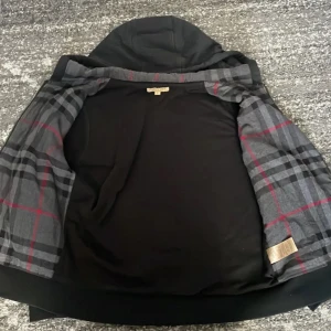 Svart hoodie från Burberry - Snygg svart hoodie från Burberry med dragkedja och huva. Insidan har ett klassiskt rutigt mönster i grått och rött. Perfekt för en stilren look. Kontakta vid frågor.