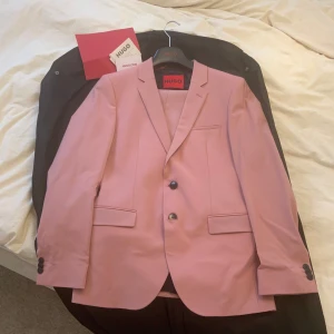 Rosa kostym från Hugo.  - Endast använd en gång! Snygg rosa kostym från Hugo med kavaj och matchande byxor. Kavajen har två knappar och klassiska slag. Perfekt för en stilren och modern look. Nypris 5000 kr. 