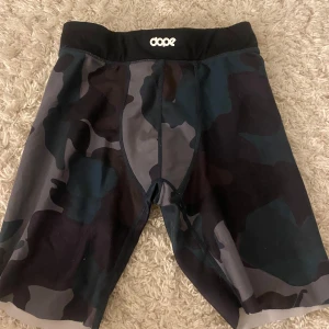 Camouflage träningsshorts från Dope - Säljer ett par coola träningsshorts från Dope i ett mörkt camouflage-mönster. Shortsen har en elastisk midja och är perfekta för intensiva träningspass. De är designade för att ge maximal rörelsefrihet och komfort.