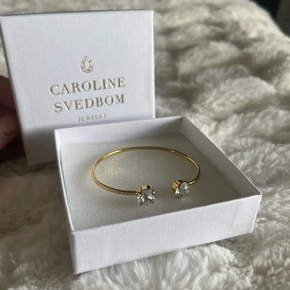 Elegant guldarmband från Caroline Svedbom med två gnistrande stenar i ändarna. Perfekt för att lägga till en touch av glamour till din outfit. Armbandet kommer i en stilren vit ask. Med en beige aktig sten . Asusteet.