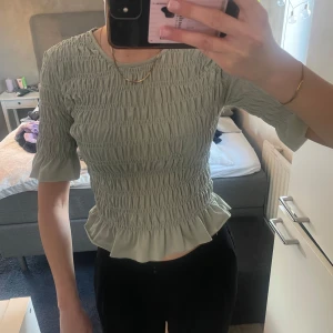 Grön Topp/tröja  - Säljer en grön topp från Gina Tricot med smockdetaljer och volang vid nederkanten. Toppen har trekvartsärmar och en rund halsringning. Perfekt för en stilren look!
